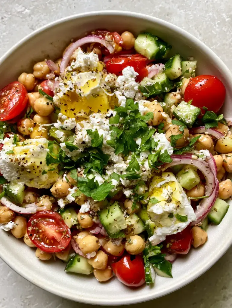 A heavy drizzle of golden lemon herb vinaigrette over a cucumber chickpea feta salad with visible oregano specks.
