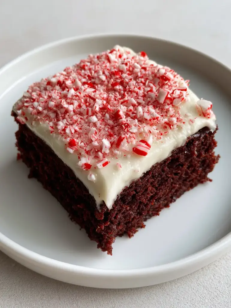 A square cut Peppermint Brownie on a white ceramic plate against a gray background, showing the texture of the white frosting and peppermint shards.