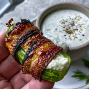 Close up of a single air-fried pickle popper held in a hand, showing the glistening charred bacon edges. (pickle poppers)
