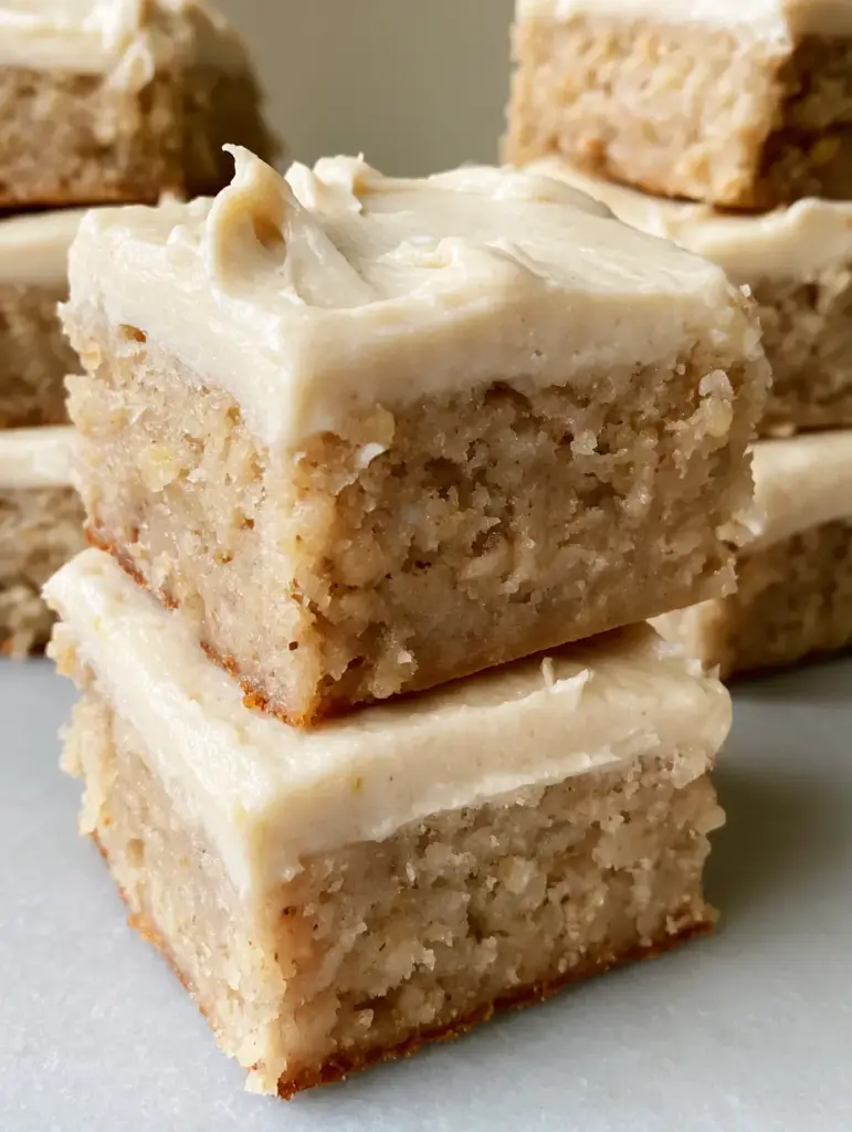 Banana Bread Brownies with Creamy Vanilla Frosting 1 Neatly stacked square slices of frosted banana bread brownies on a neutral gray surface.