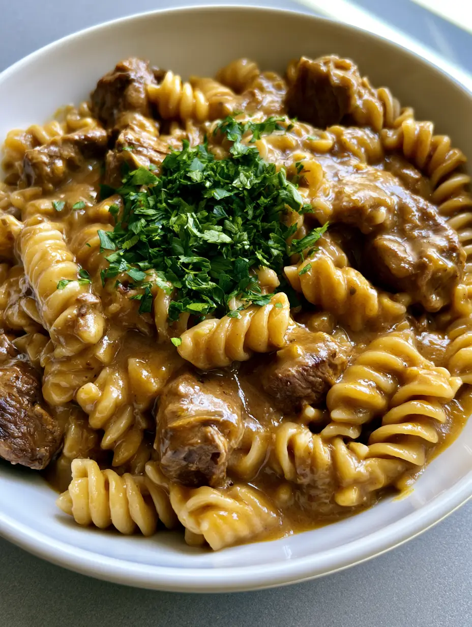 Slow Cooker Garlic Beef Pasta with Creamy Sauce 4 A handheld smartphone photo of a white ceramic bowl filled with cooked spiral rotini pasta and tender beef tips in a light-brown garlic sauce. (Slow Cooker Garlic Beef Pasta)